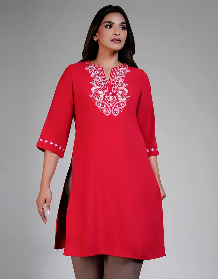 A red tunic with white embroidered floral design on the chest, V-neck, and 3/4 sleeves with embroidered cuffs. Paired with neutral-colored pants. Model on gray background.