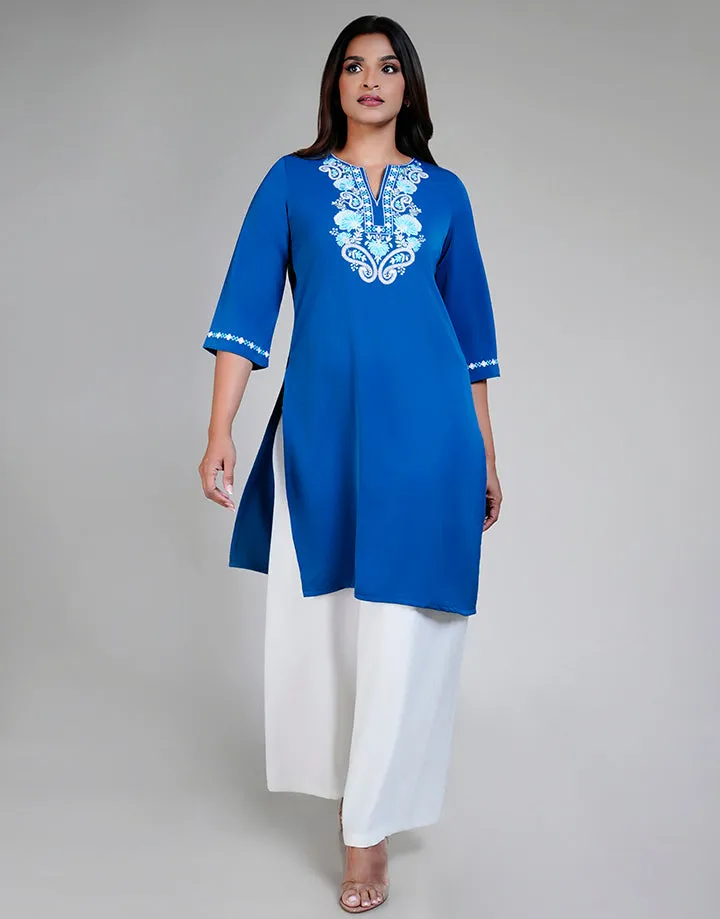 A blue tunic with white embroidered floral design on the chest and 3/4 sleeves with embroidered cuffs. Worn over white wide-leg trousers. V-neck, side slits. Model on gray background.