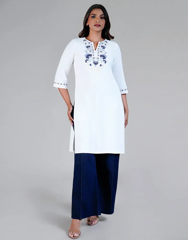 A white tunic with blue embroidered design on the chest and cuffs, featuring a V-neck and 3/4 sleeves. Styled over dark wide-leg pants. Model on gray background.