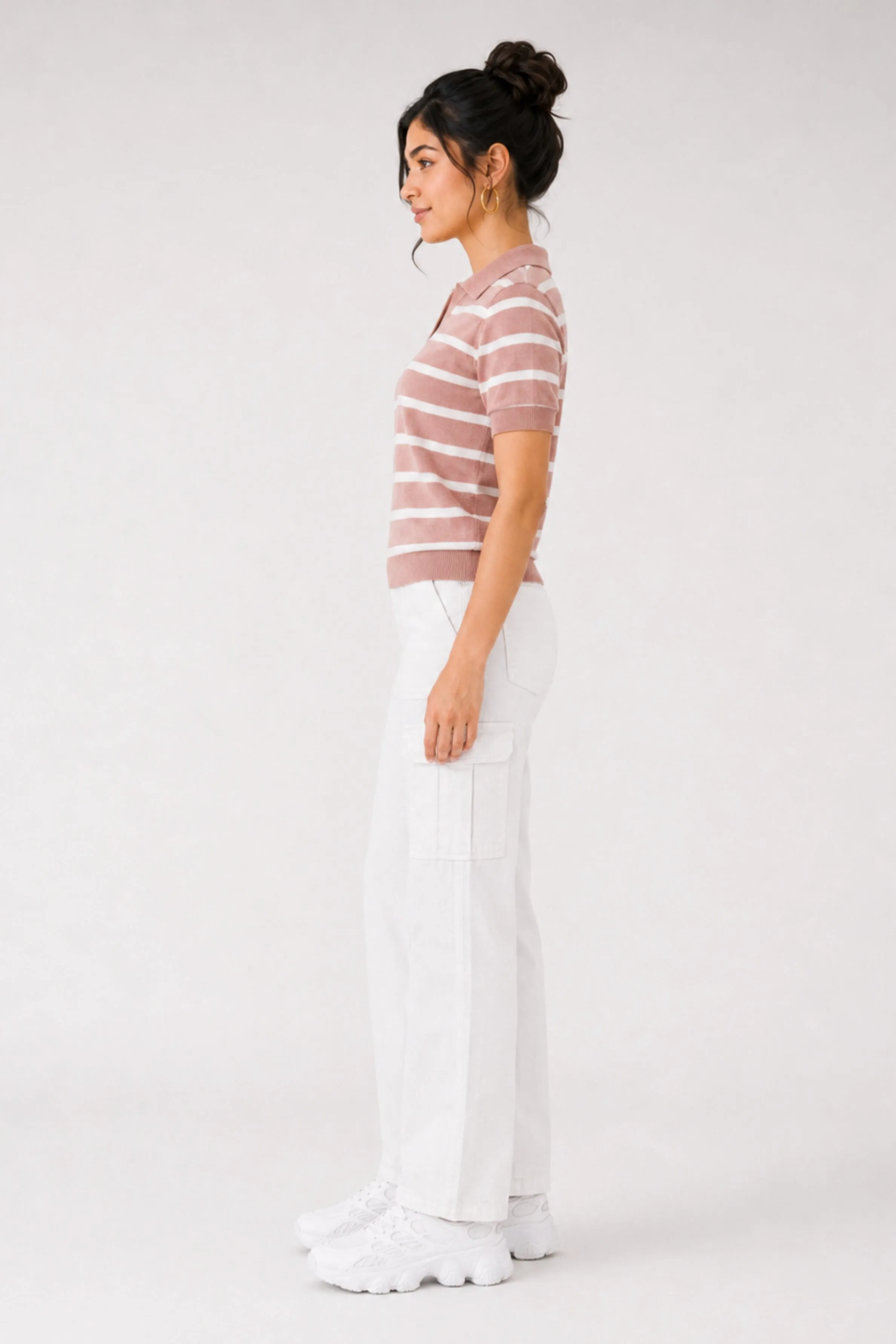 A side view of a model wearing a pink striped polo shirt with short sleeves and white cargo pants, accessorized with white sneakers, against a plain background.