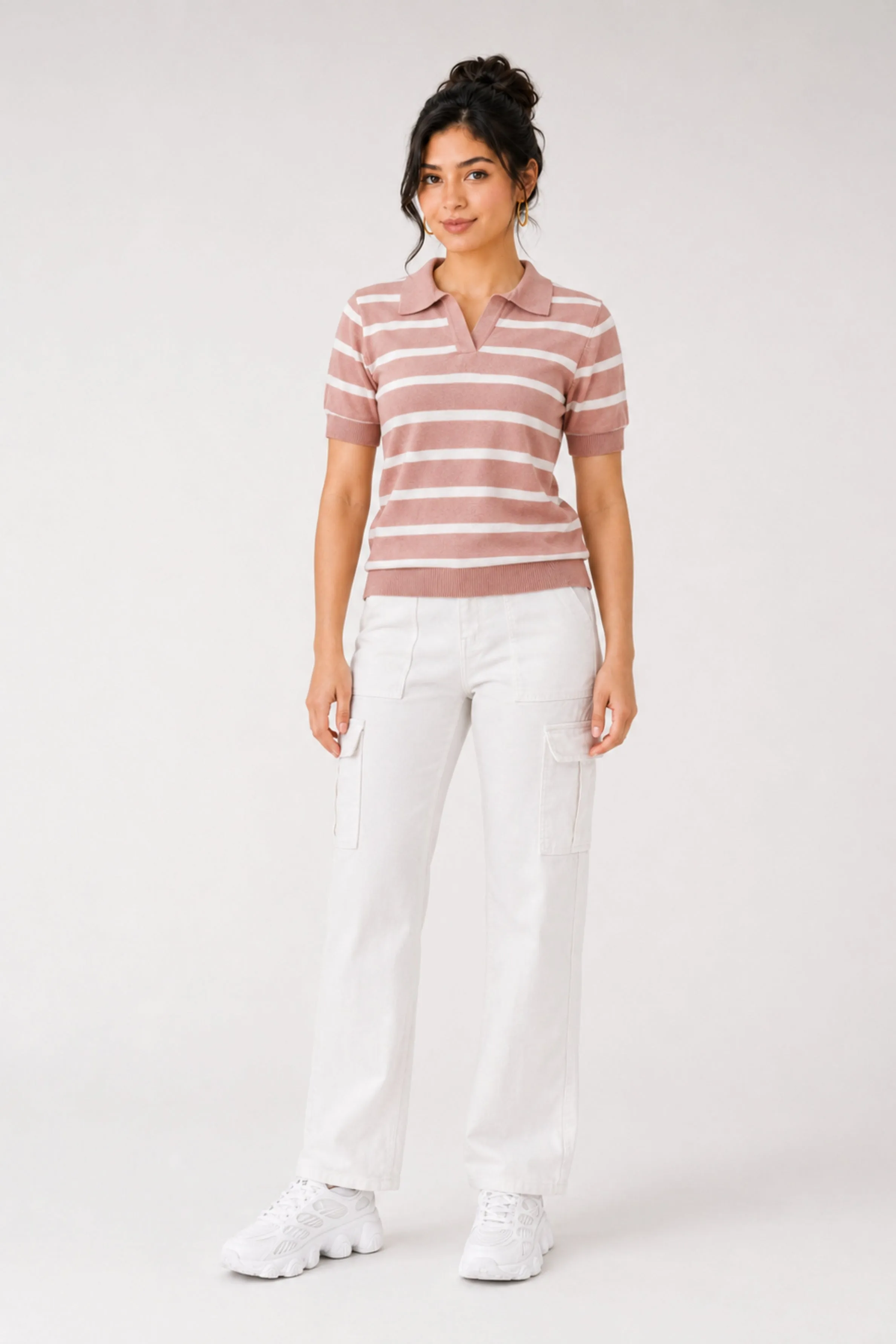 A model wears a pink and white striped knit polo shirt with short sleeves, paired with white cargo pants and white sneakers. The outfit is styled against a plain background.