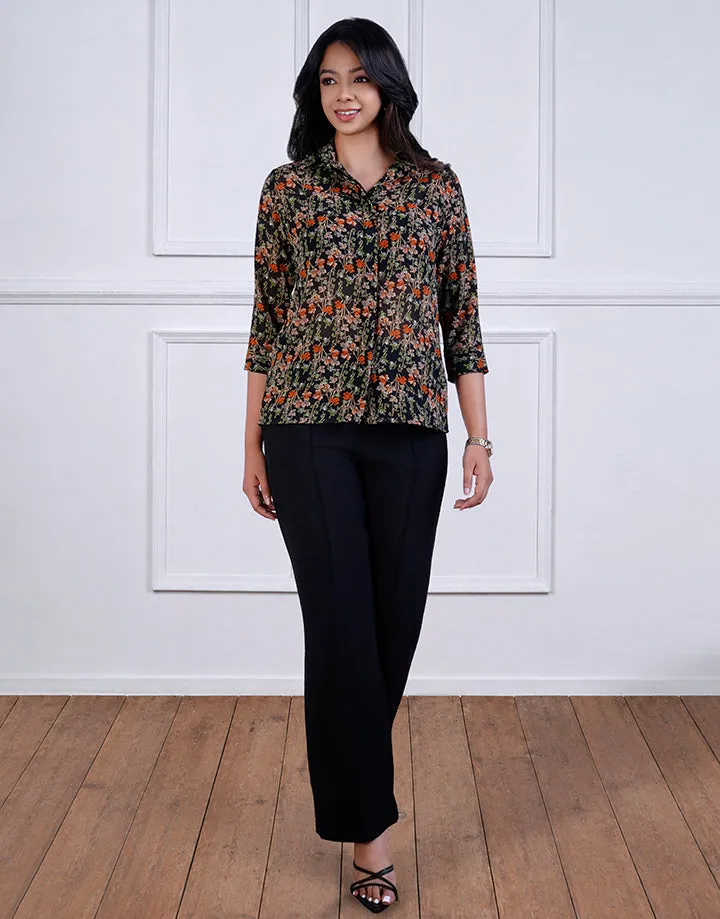 A woman wearing a floral-patterned blouse with black pants. The blouse has a collar and three-quarter sleeves. She's standing on a wooden floor against a white paneled wall.