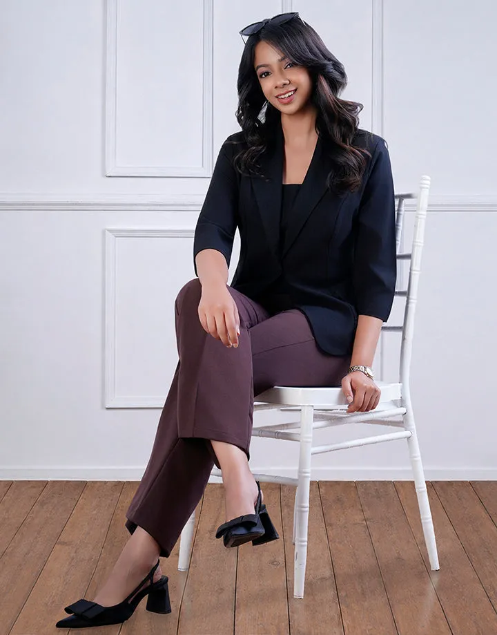 A woman seated wearing a black blazer over a black top with brown pants. She has on black heels with bows. The setting includes a white chair on a wooden floor with a white backdrop.