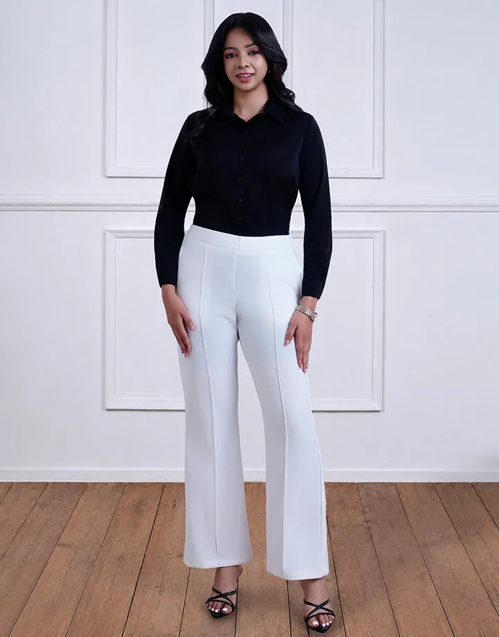 A woman wearing a black button-down shirt and white pants. She's standing on a wooden floor with a white paneled wall backdrop, facing the camera.
