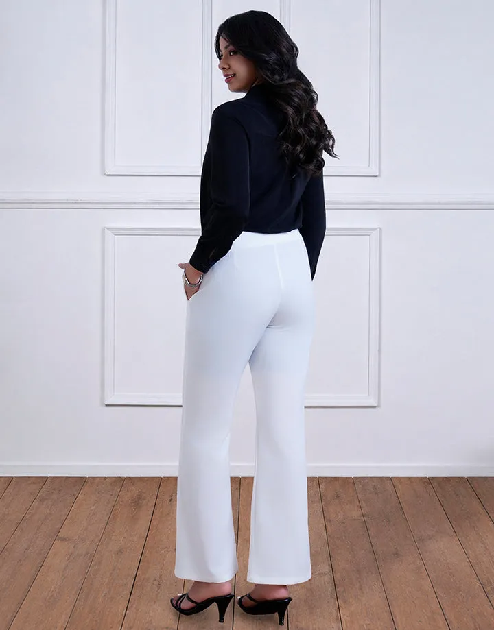 A woman in a black top and white high-waisted pants. She is standing on a wooden floor, facing away from the camera. The backdrop is a white paneled wall.