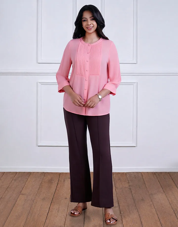 A woman wearing a pink blouse with button details and three-quarter sleeves, paired with brown pants. She stands on a wooden floor against a white paneled wall.