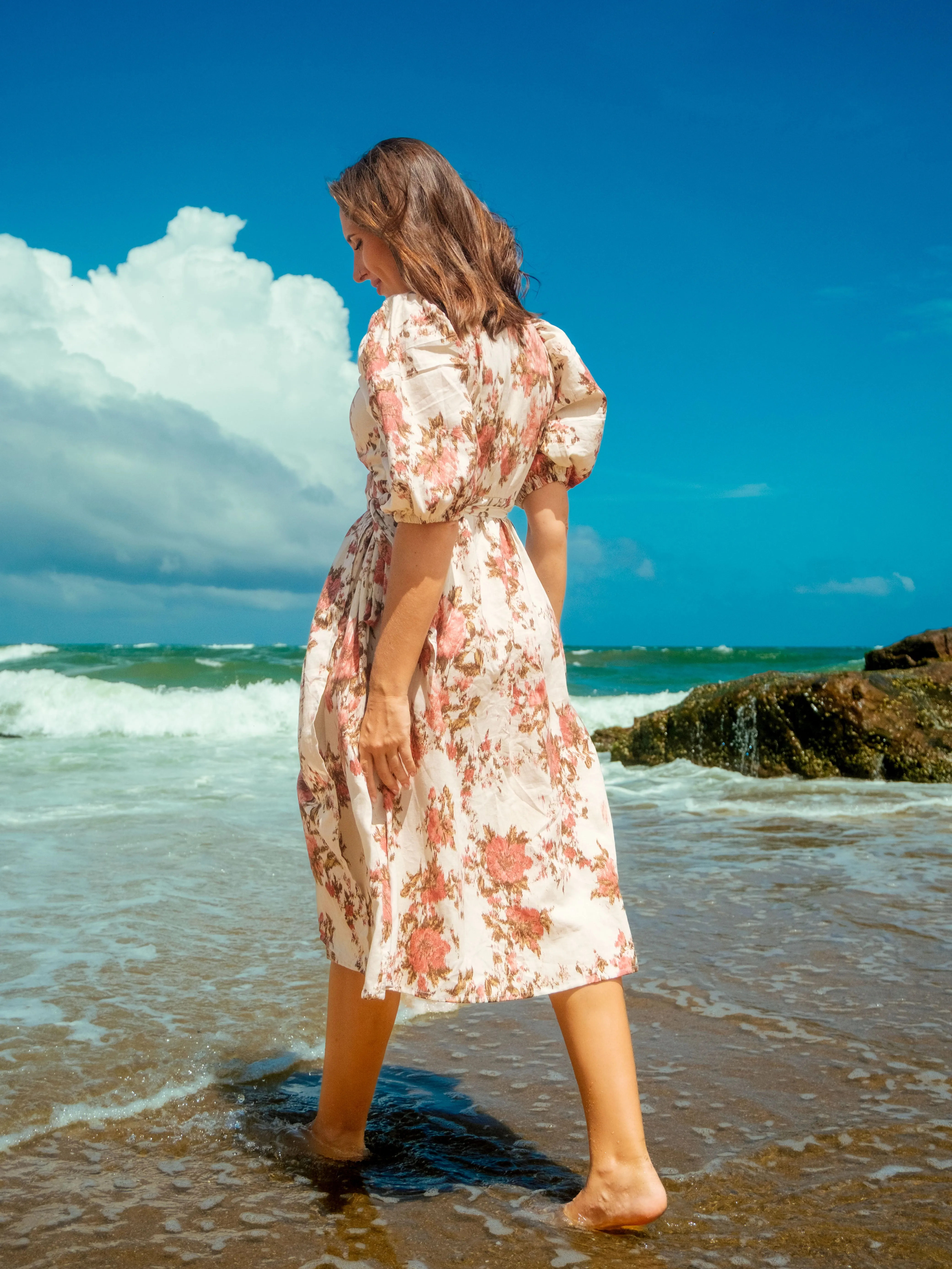 Back view of woman wearing floral midi dress with puff sleeves, gathered waist on beach. Dress is white with pink floral patterns. She walks barefoot in shallow water. Ocean waves and bright blue sky.