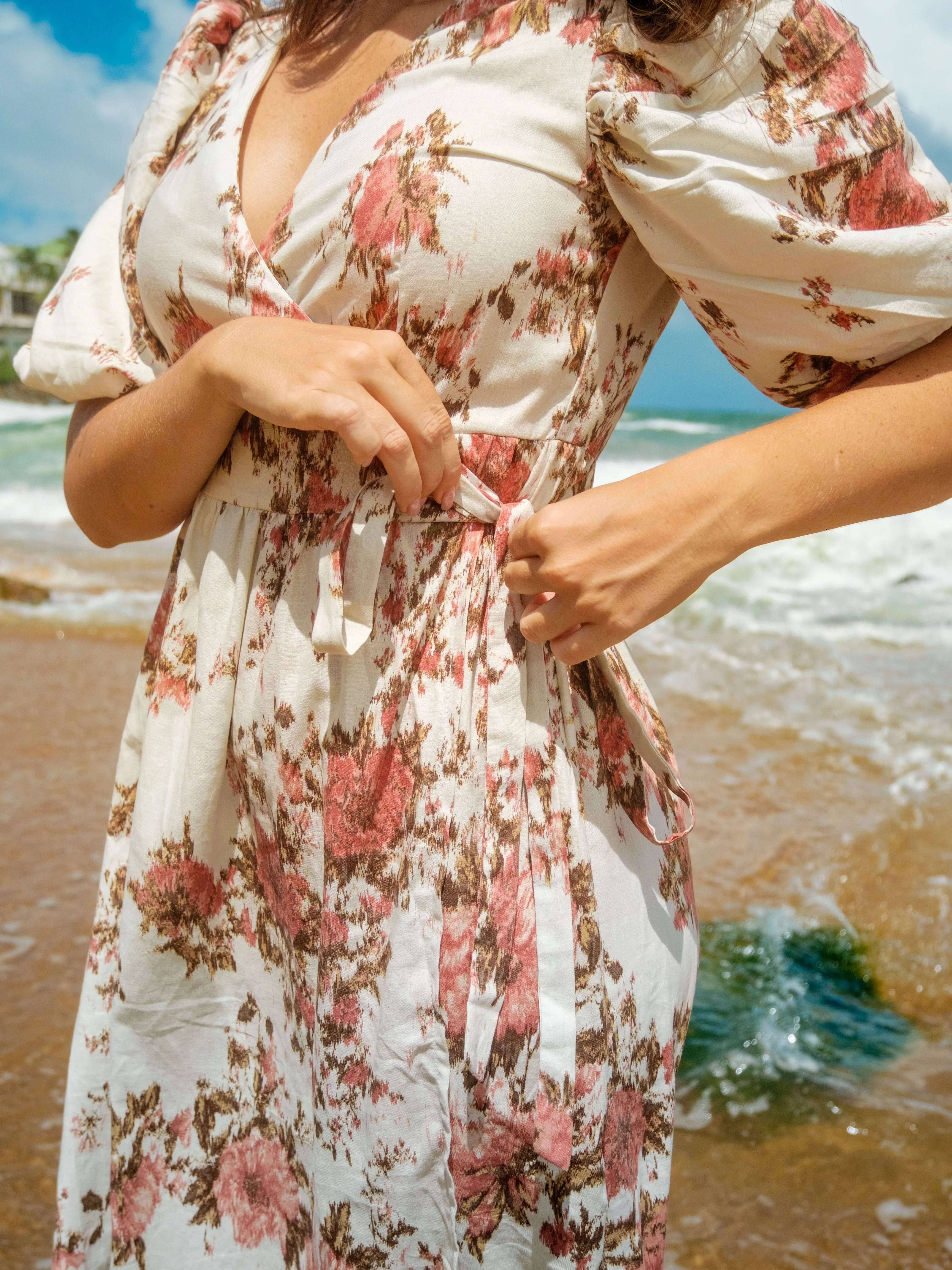 Close-up of floral wrap dress with tie detail at waist. Dress is white with pink floral pattern, features V-neck, puff sleeves. Model adjusts tie, stands on beach. Ocean visible in background.