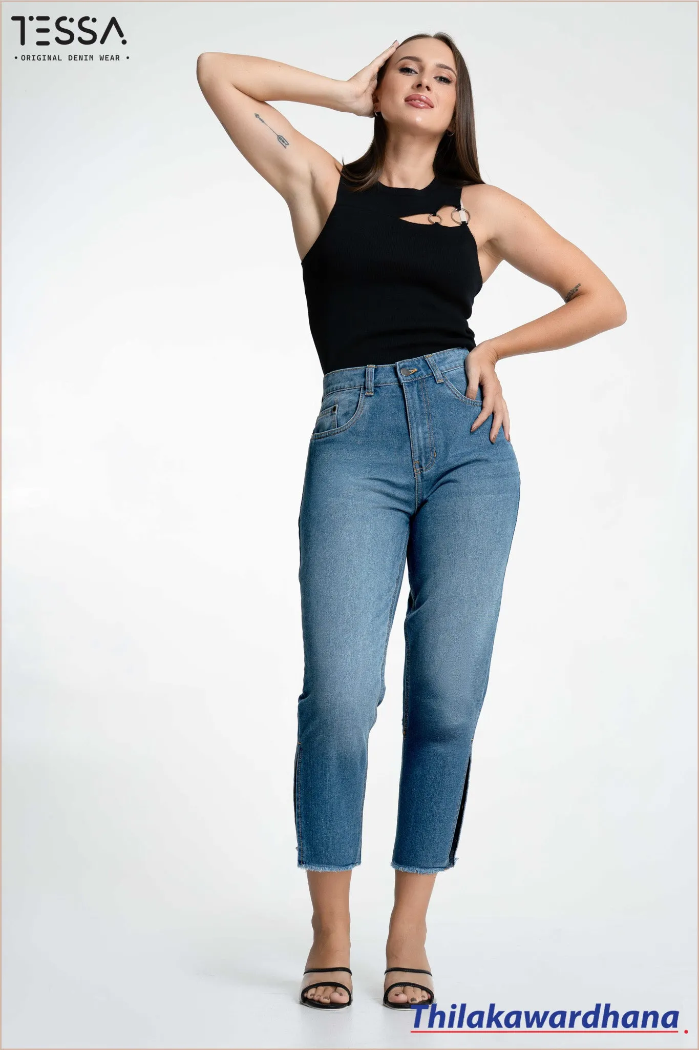 Model posing in a black sleeveless top with metallic ring detail, fitted blue jeans with a cropped and slit design. Styled with black sandals. Background is white, with TESSA logo and Thilakawardhana branding.