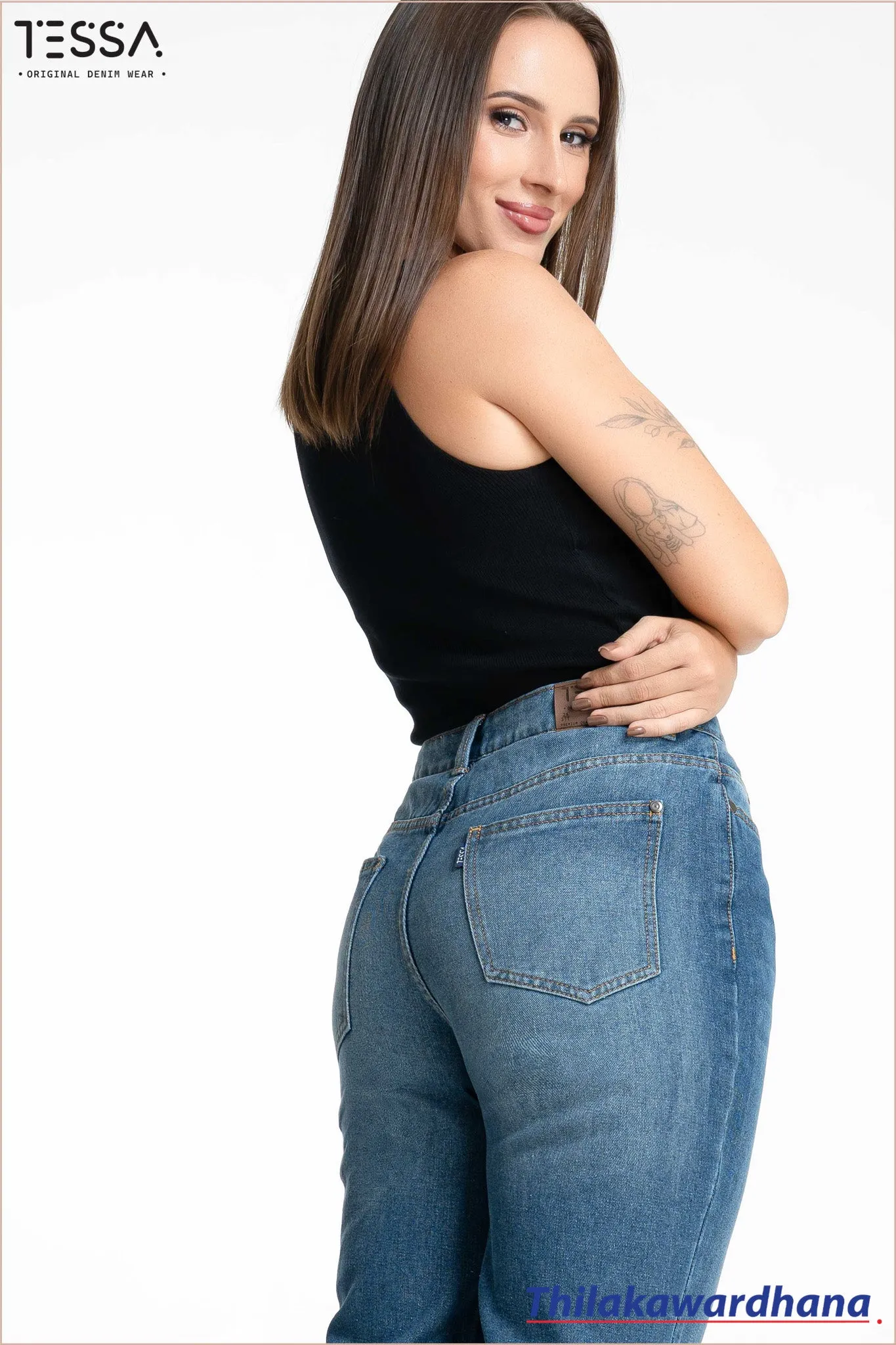 Close-up of model in a black top and blue jeans, showing back pocket details and top's ring accent. Model has a tattoo on upper arm. Background is plain white with TESSA logo at top left and Thilakawardhana at bottom right.