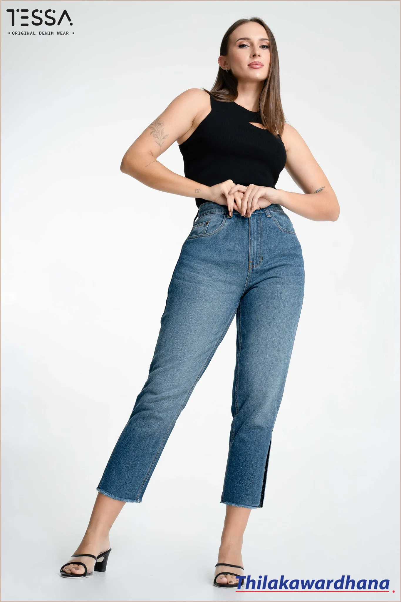Model in black sleeveless top featuring a single shoulder metallic ring, with fitted blue jeans. Barefoot with hands on waist. Plain white background with TESSA and Thilakawardhana branding.