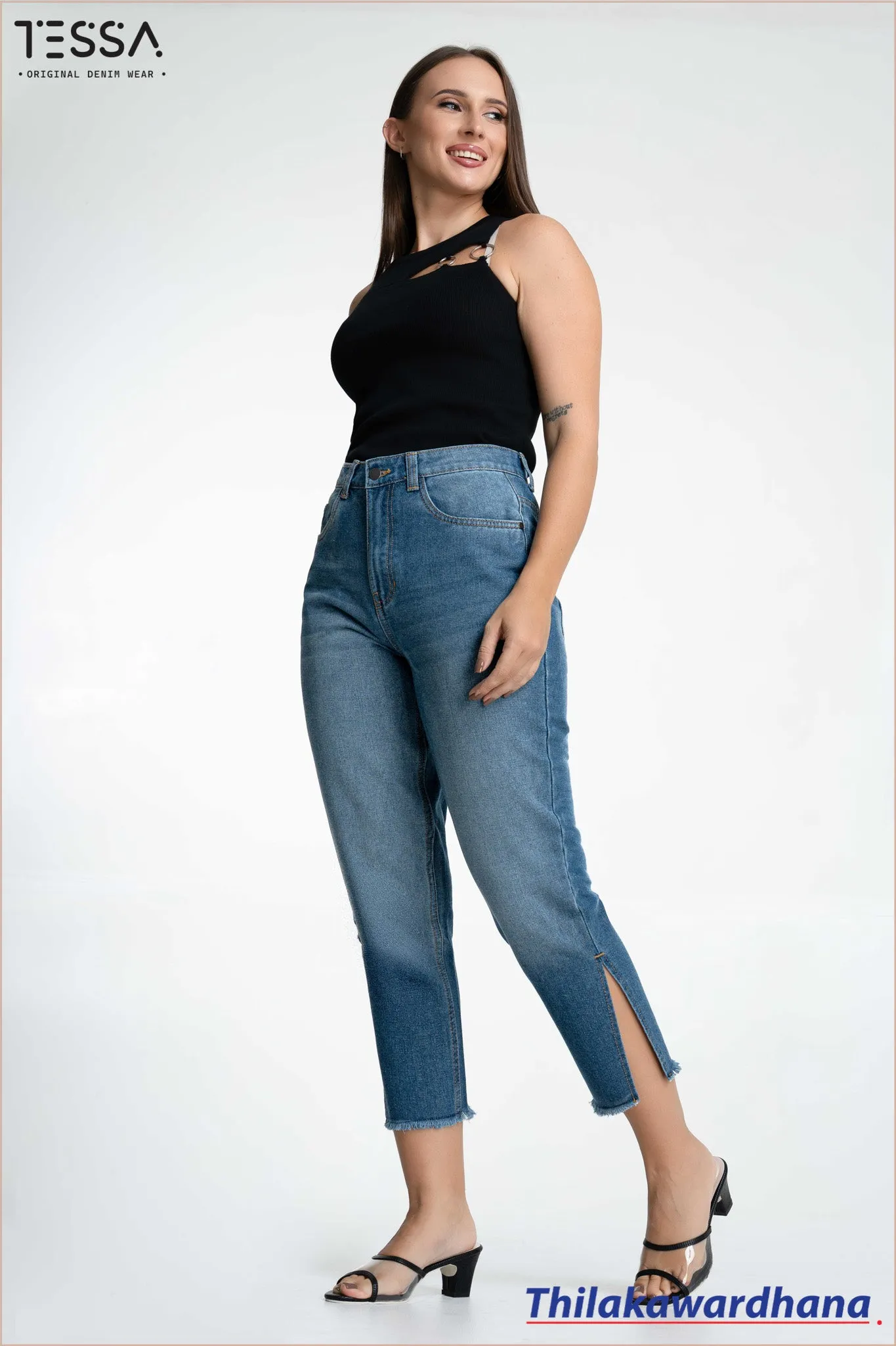 Model wearing a black sleeveless top with metallic ring detail on the shoulder, paired with high-waisted blue jeans, cropped with side slits. Background is plain white. Visible TESSA logo at the top left and Thilakawardhana at the bottom right.
