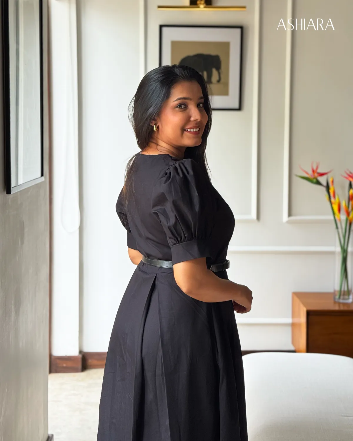 A profile view of a woman in a black puff-sleeve dress with buttons and a belt, photographed indoors. The decor includes floral elements and framed art. Visible branding: ASHIARA.