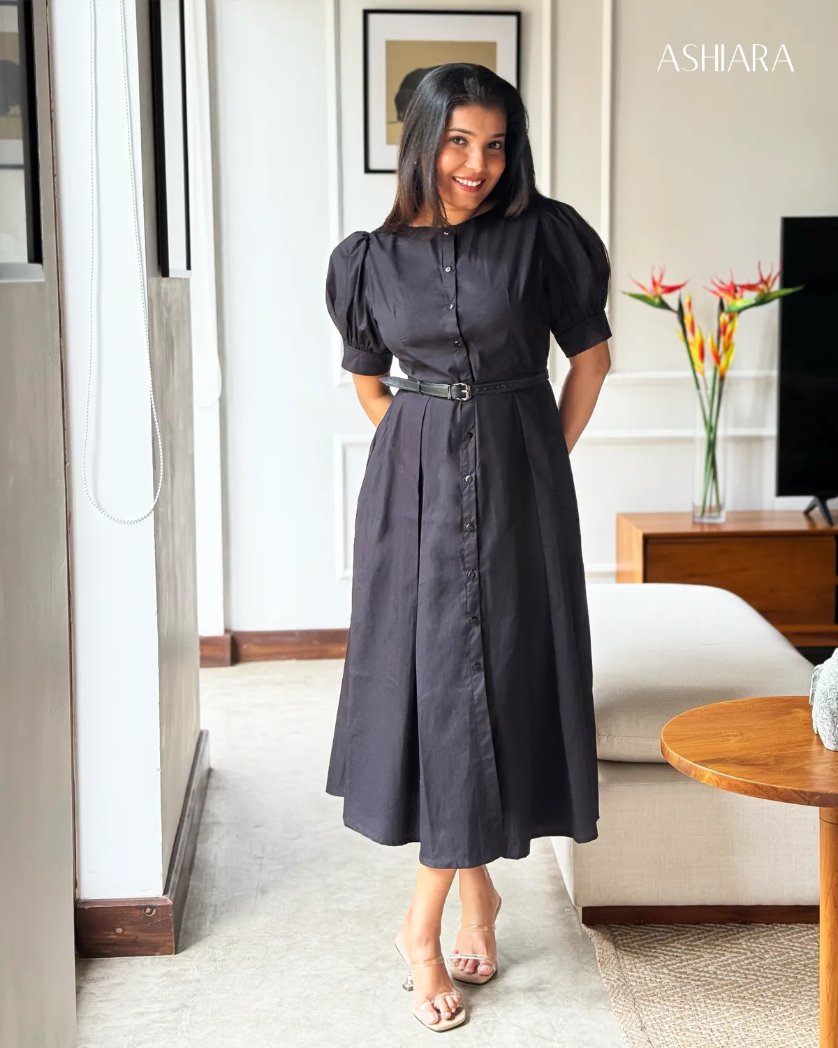 A full-length view of a black button-down dress with puff sleeves and a waist belt, worn by a woman standing indoors. The setting includes a modern living space with wall art. Visible branding: ASHIARA.